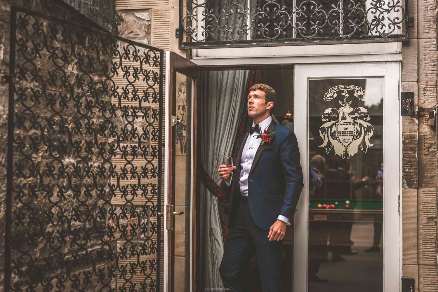 Groom in navy suit with burgundy bow tie standing in ornate doorway of elegant wedding venue.