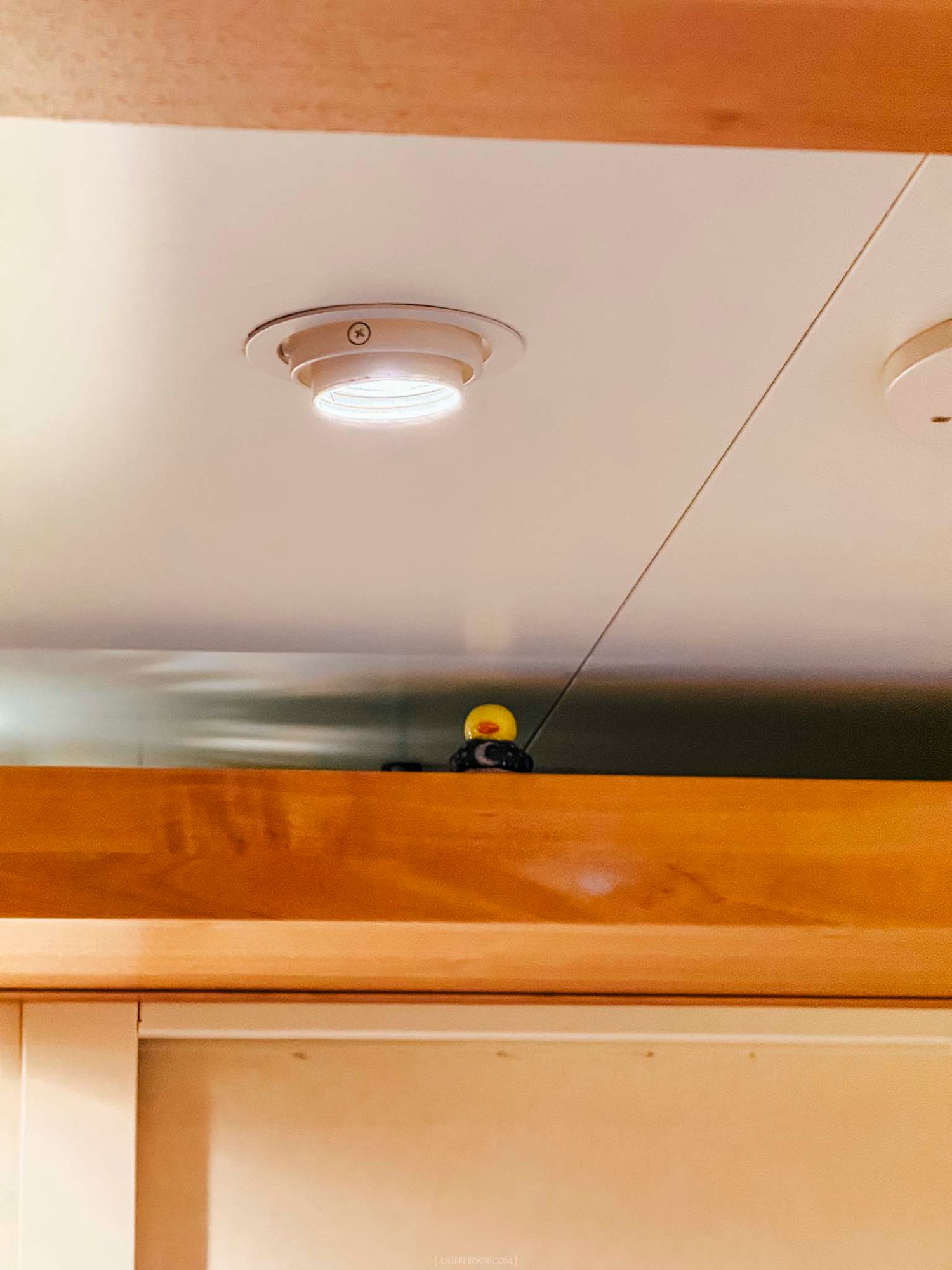 White LED puck light mounted on a ceiling panel above wooden cabinet shelving in a modern interior space.