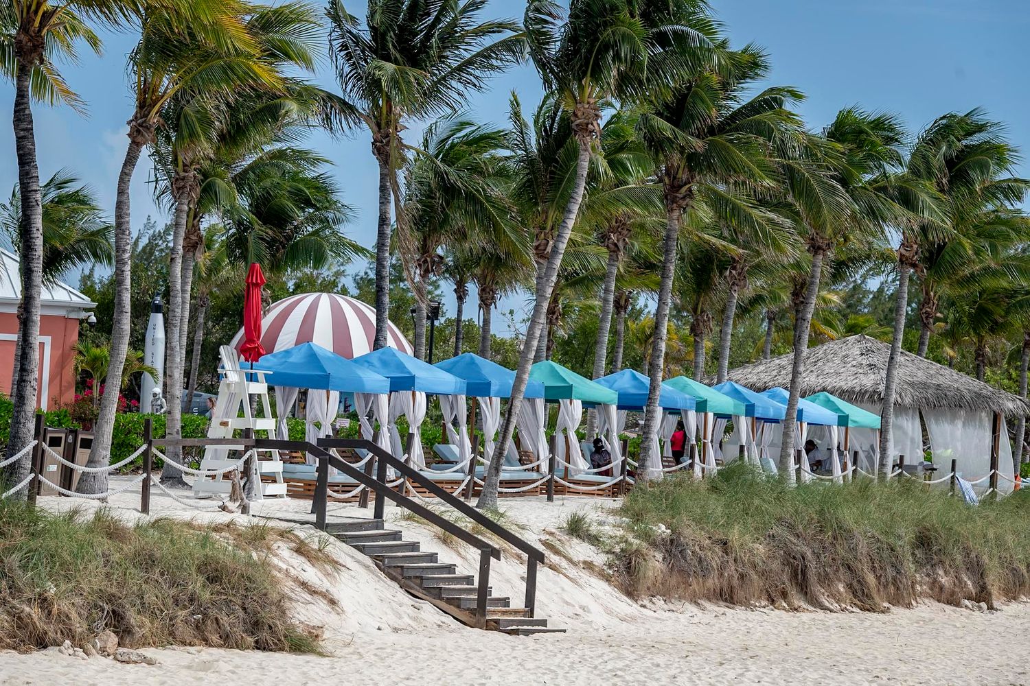 Tropical beach resort with colorful umbrellas, palm trees swaying in wind, and thatched huts on white sandy shore.