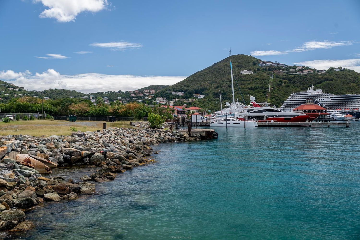 Rocky shoreline with yachts docked at a marina, lush green hills, and a cruise ship in a tropical Caribbean harbor.