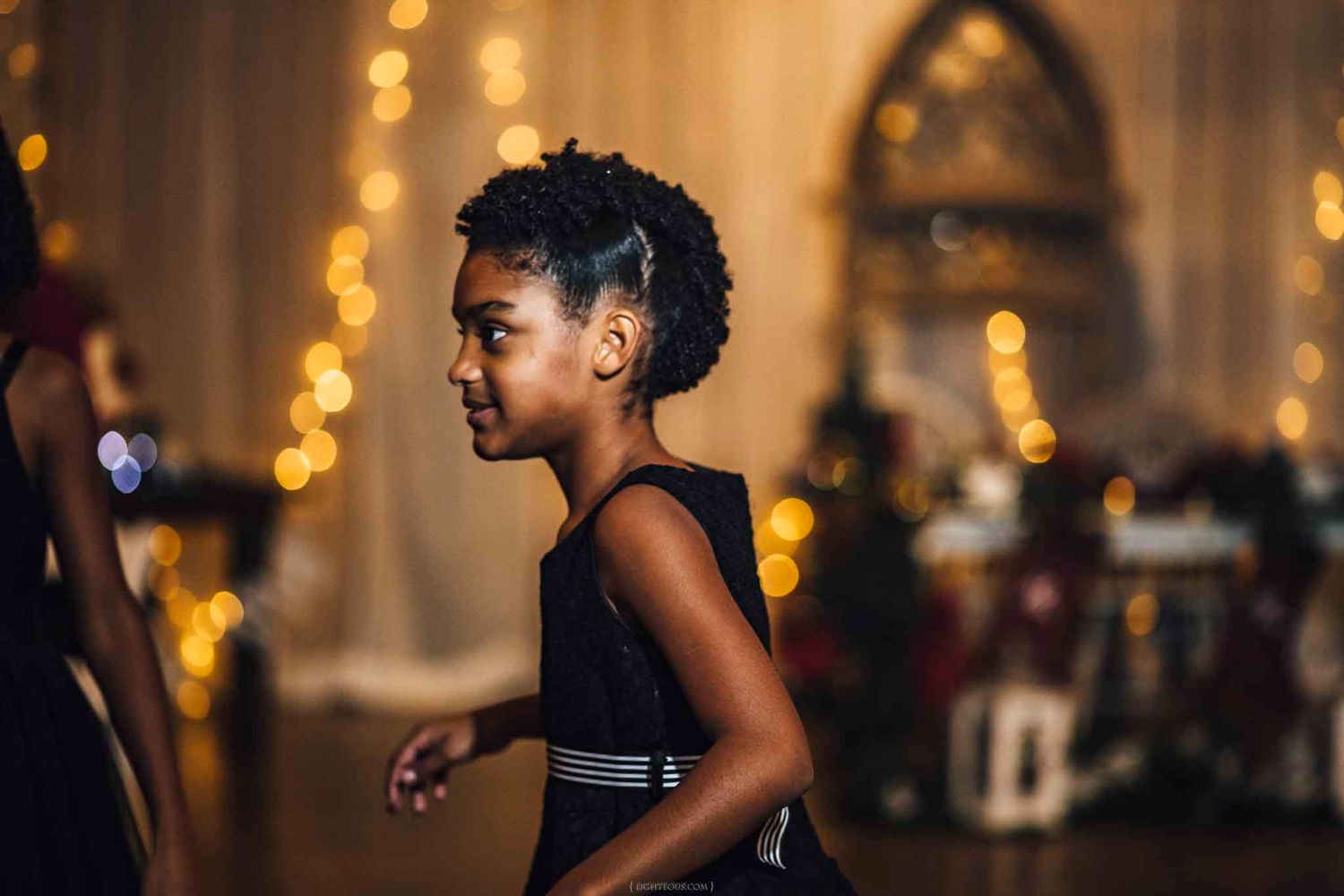 Young Black girl in a sleeveless dress dancing at a festive event with warm bokeh lights in the background.