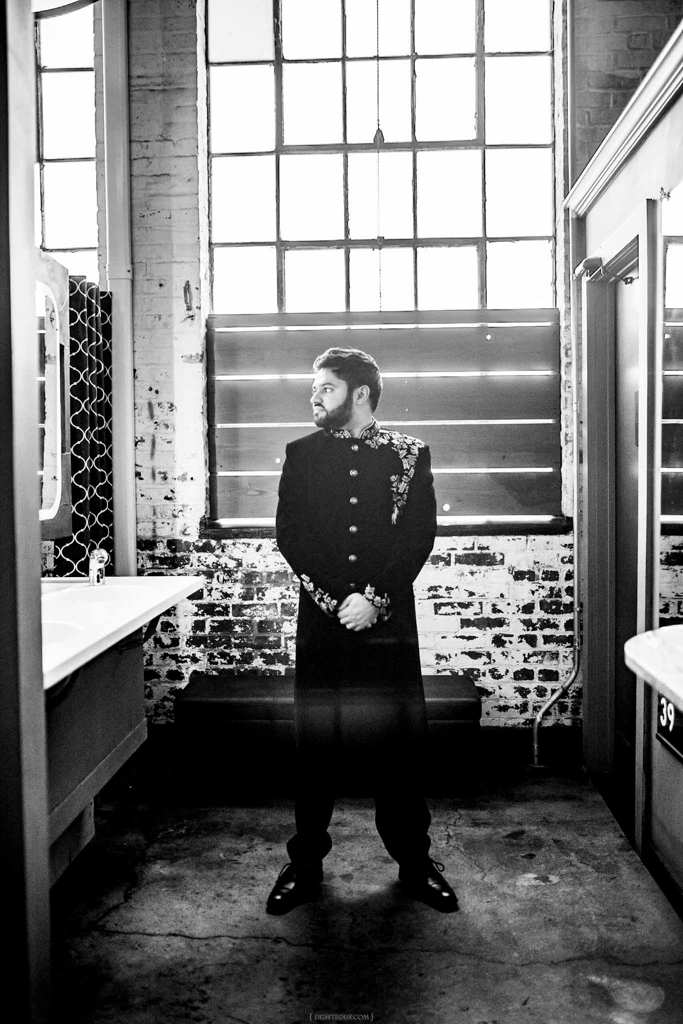 Black and white photo of a bearded man in traditional South Asian attire standing before a large industrial window.