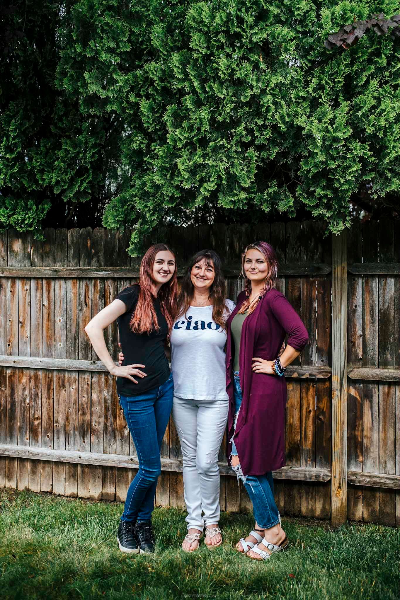Three women smiling and posing together in a backyard with a wooden fence and green tree.