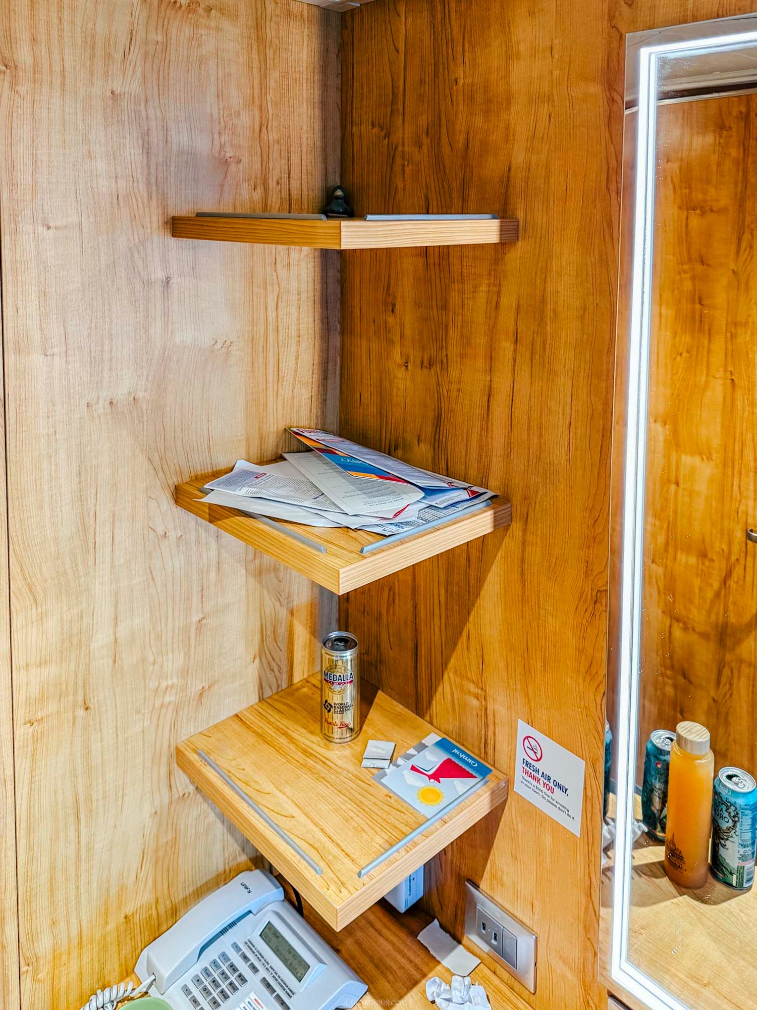 Wooden corner shelves displaying newspapers, a phone, and miscellaneous items next to a mirror in a cozy interior.