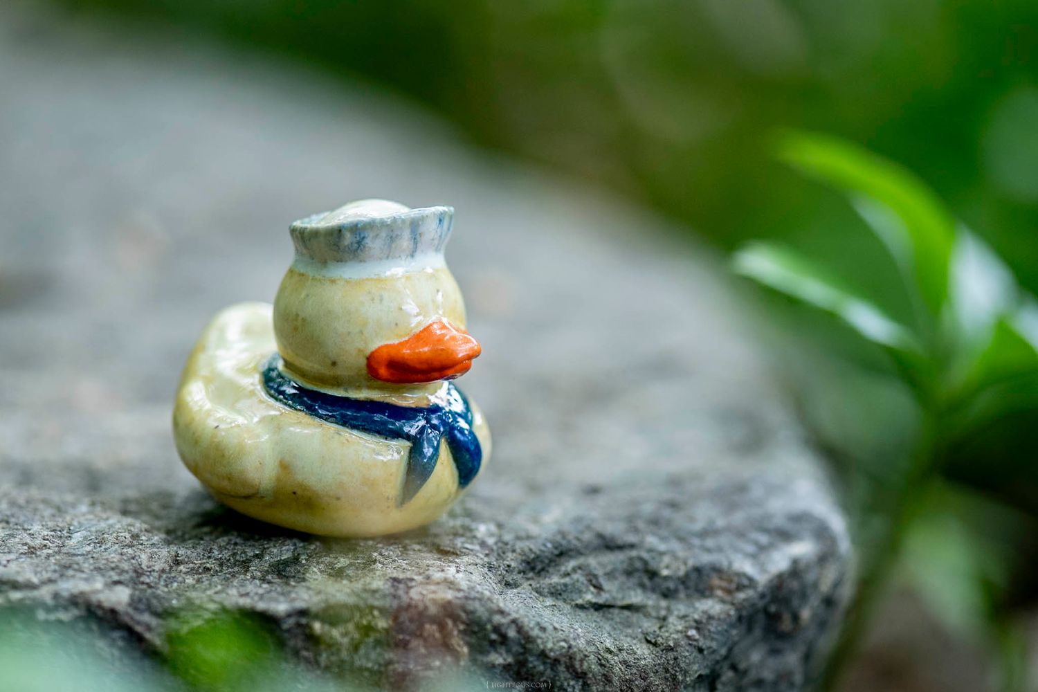 Miniature ceramic duck figurine wearing a blue scarf and white hat on a rocky surface.