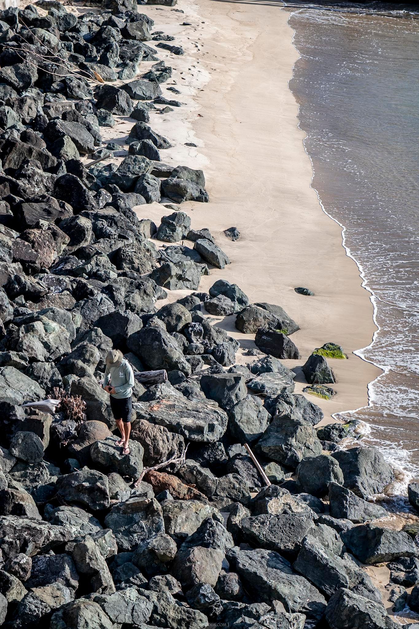 Person walking on rocky shoreline beside a sandy beach with gentle ocean waves in warm sunlight.