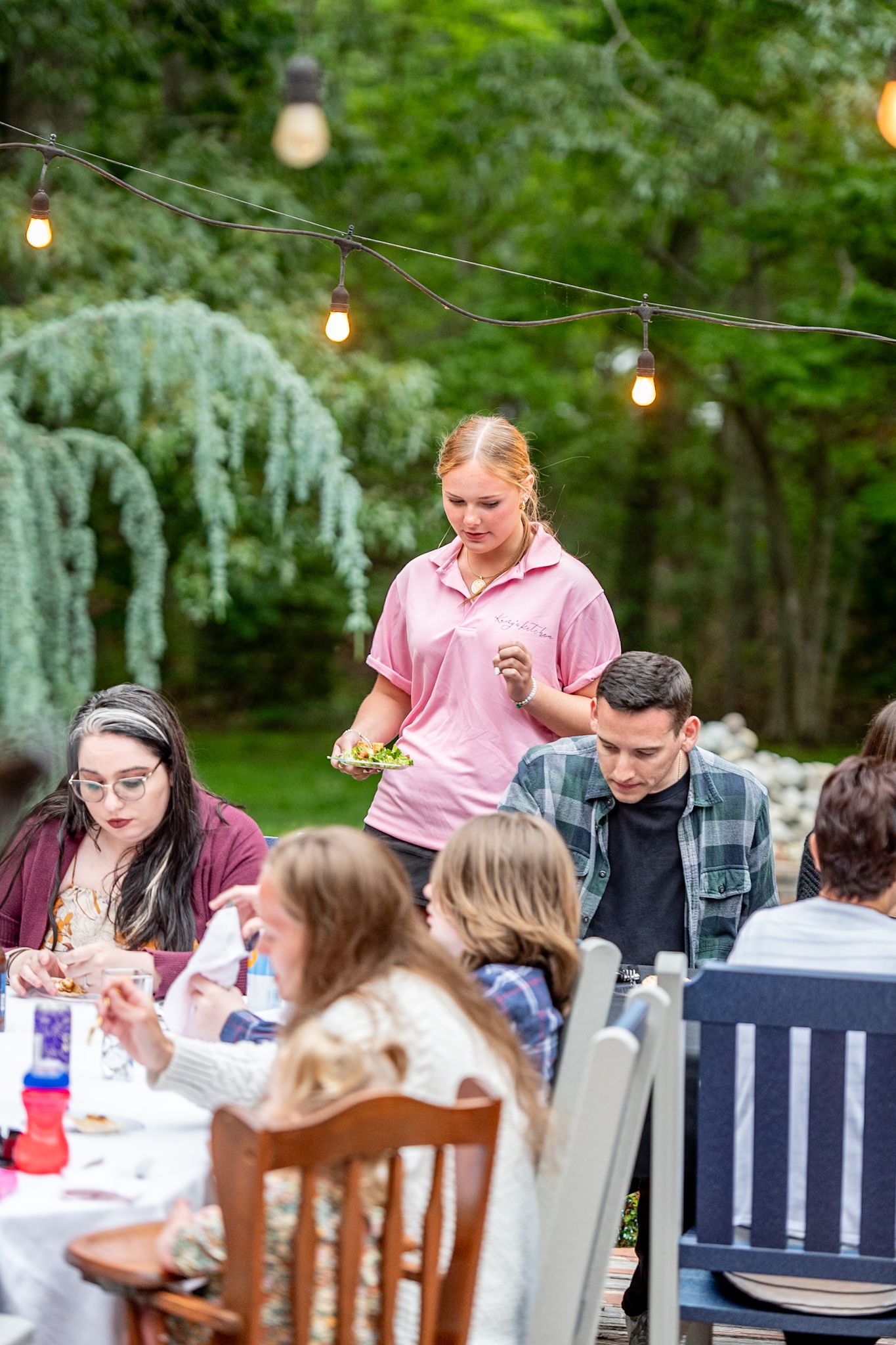 Outdoor dinner party setup with string lights and guests seated at a long table.