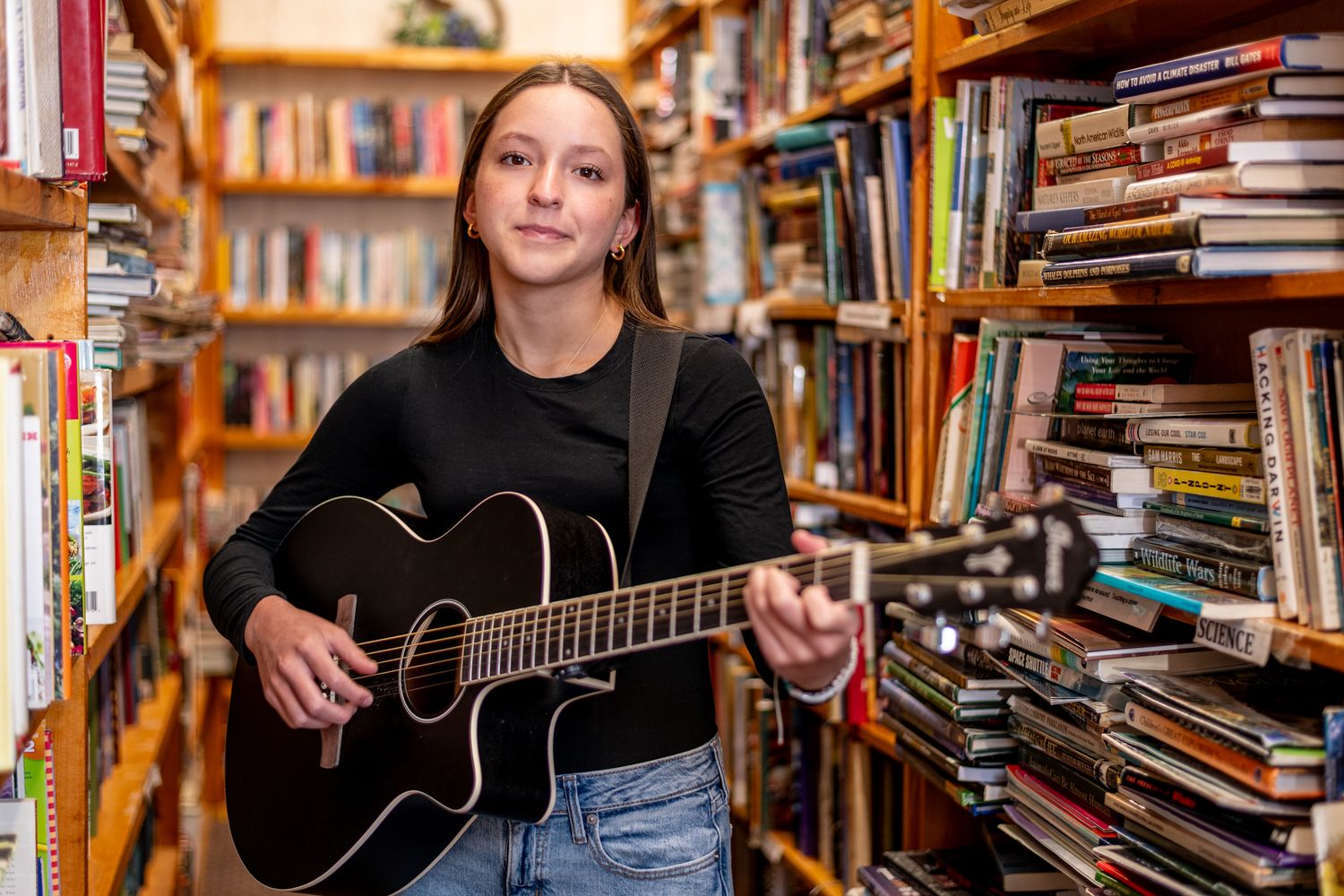 A person plays acoustic guitar while sitting among floor-to-ceiling bookshelves in a library setting.