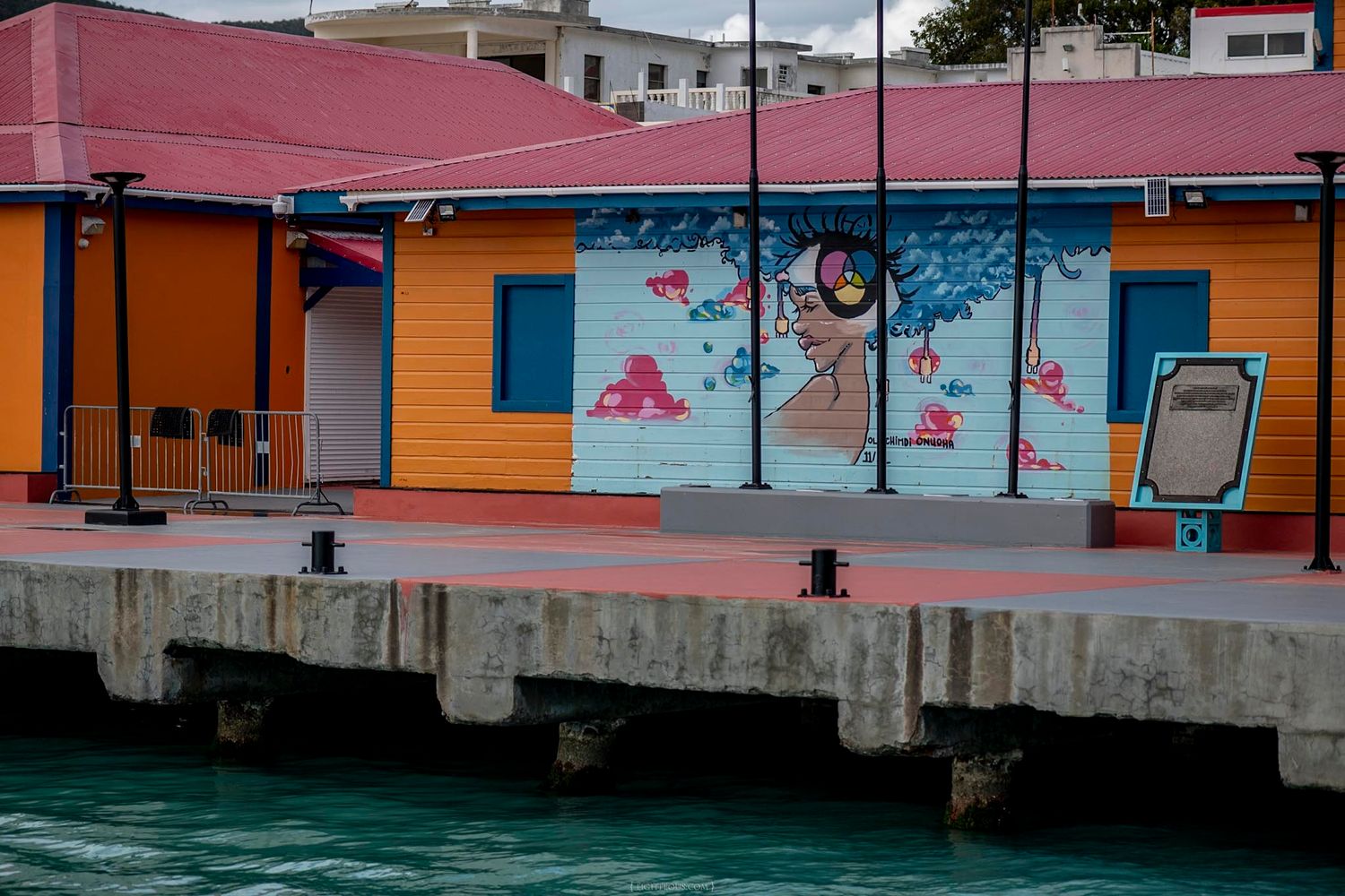 Colorful waterfront building with vibrant mural art on a Caribbean dock, featuring orange and pink facades near turquoise water.