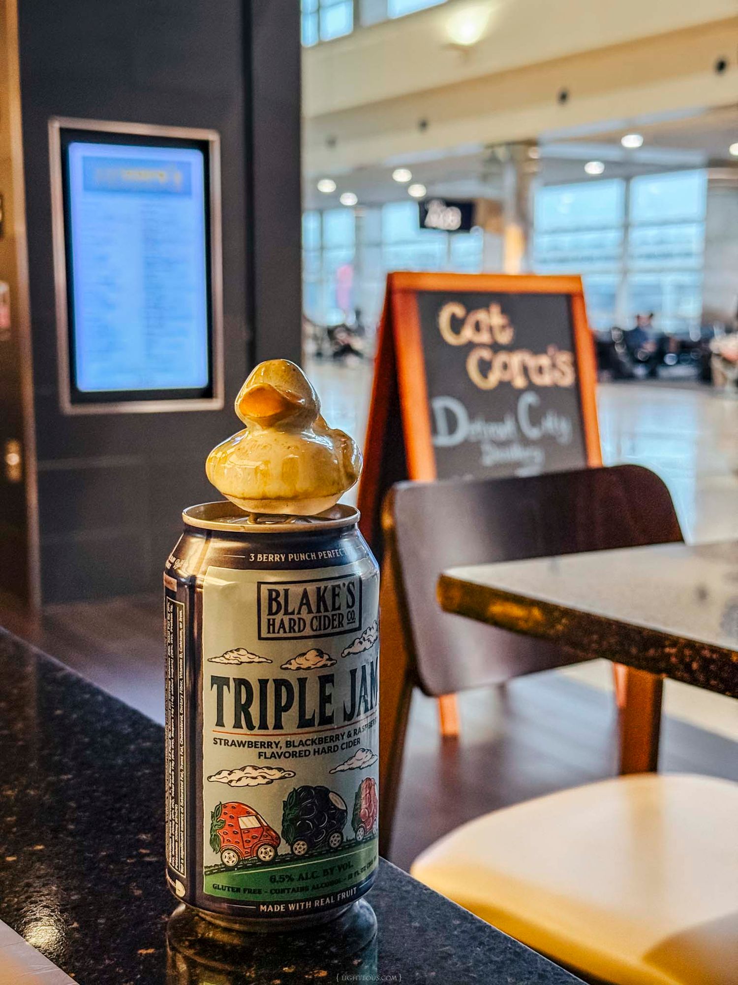 A can of Blakes Hard Cider Triple Jam sits on a bar counter with a rubber duck on top inside an airport terminal.