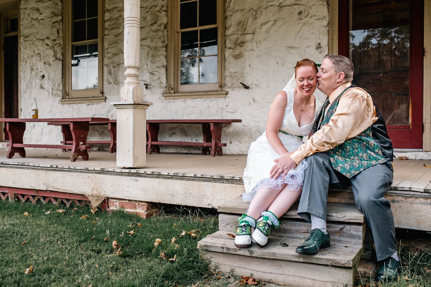 Couple in wedding attire sitting on porch steps of rustic building, bride wearing colorful sneakers, sharing a joyful moment.