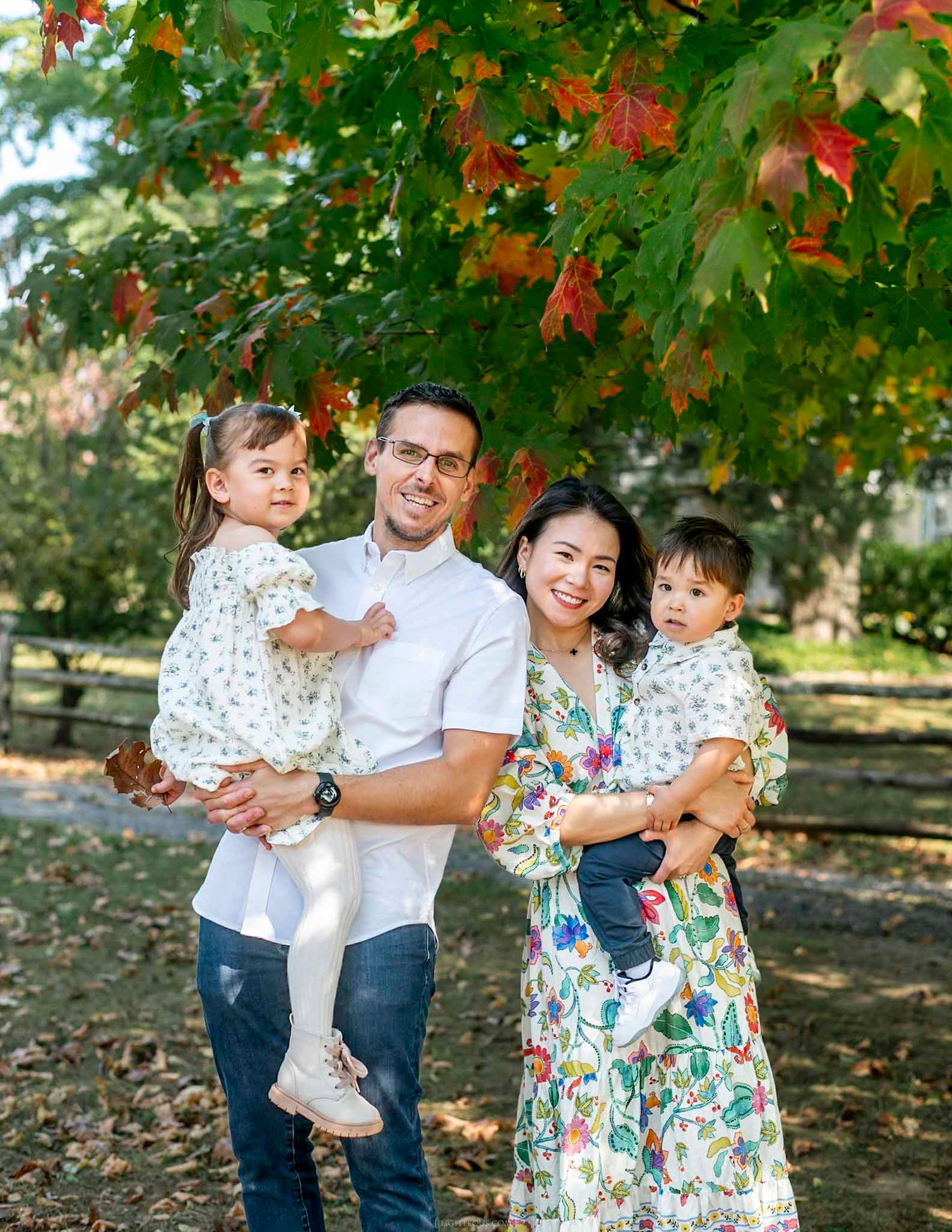 Family of four posing outdoors under colorful autumn maple tree, parents holding two young children.