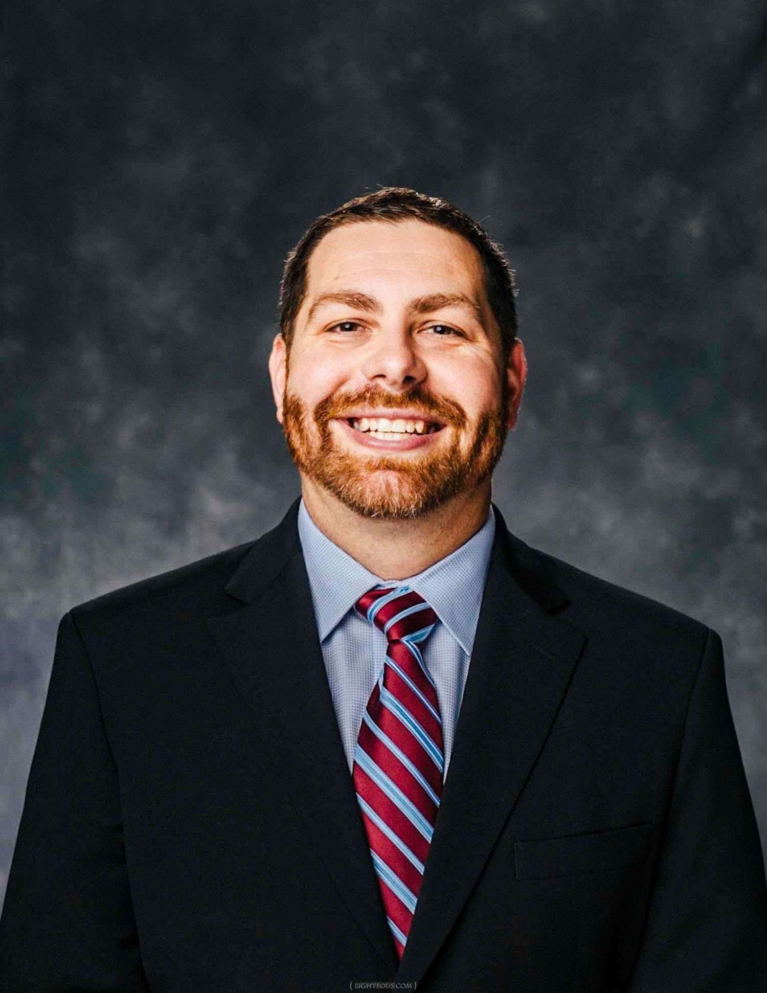 Professional headshot of a smiling man with beard wearing a dark suit and striped tie against gray background.