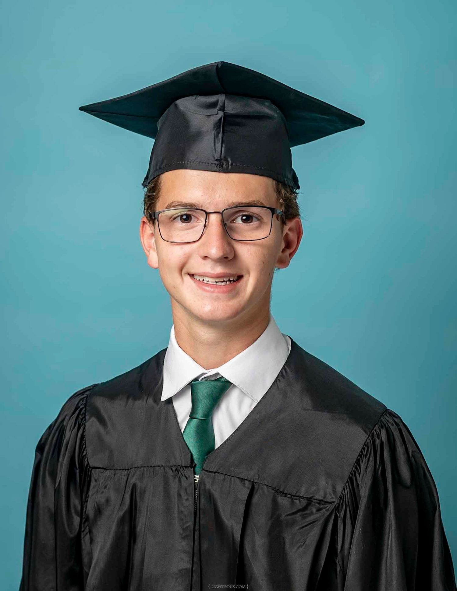 Smiling young graduate in black cap and gown with green tie and glasses posing for official graduation portrait.