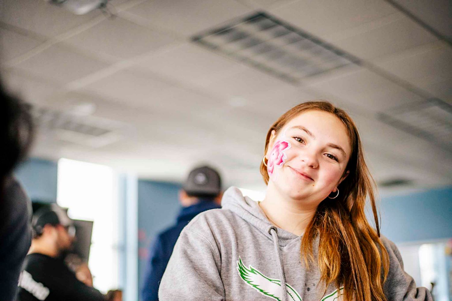 Smiling teenage girl with face paint on her cheek wearing a gray hoodie in a bright indoor setting.