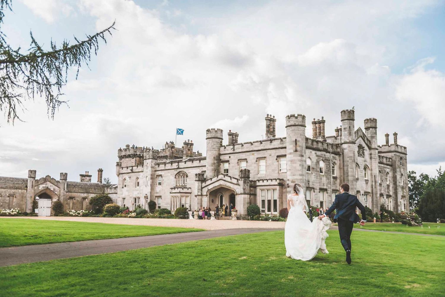 Bride and groom walking hand in hand across lush green lawn toward a grand historic castle wedding venue.