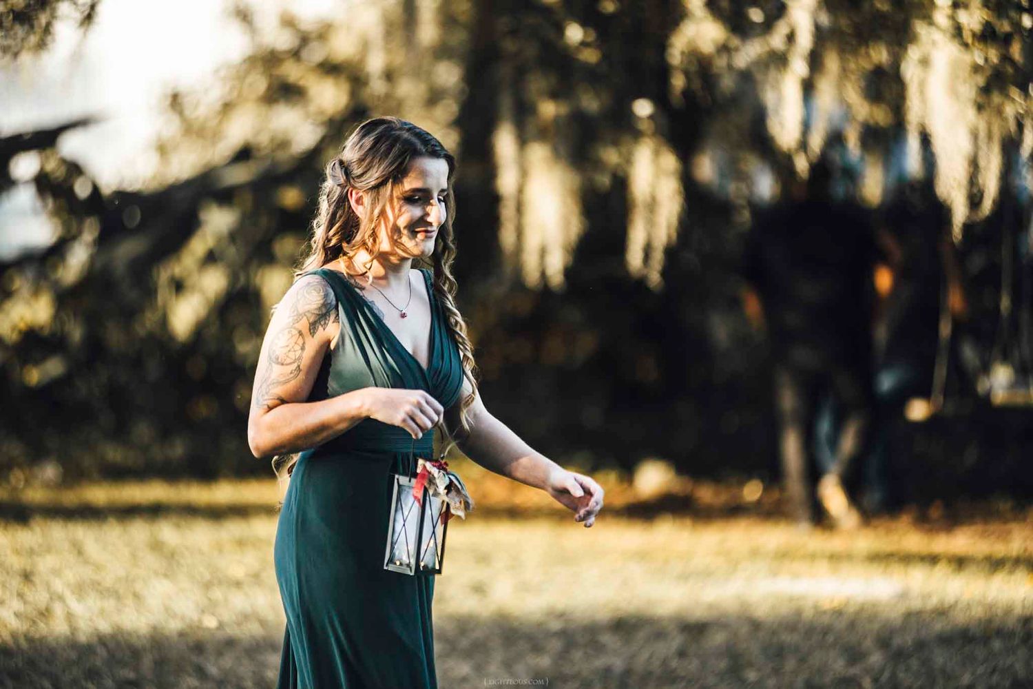 Woman in green dress holding a lantern outdoors among moss-draped trees in golden light.