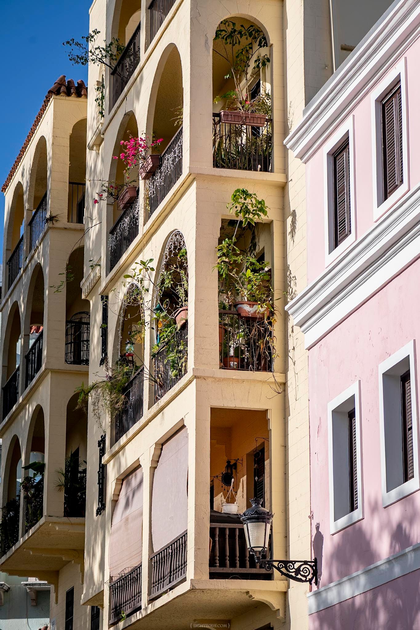 Colorful Mediterranean building facade with arched balconies adorned with lush green plants and pink flowers under a clear blue sky.