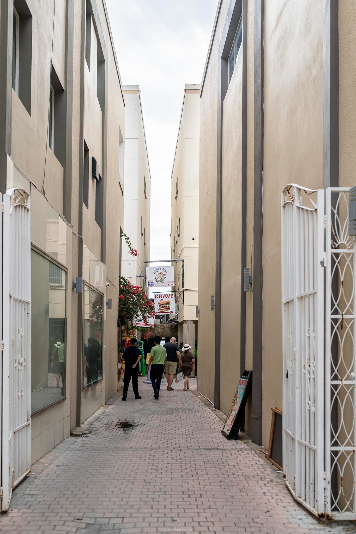 Narrow alleyway between beige buildings with people walking toward a market sign, paved with brick tiles.