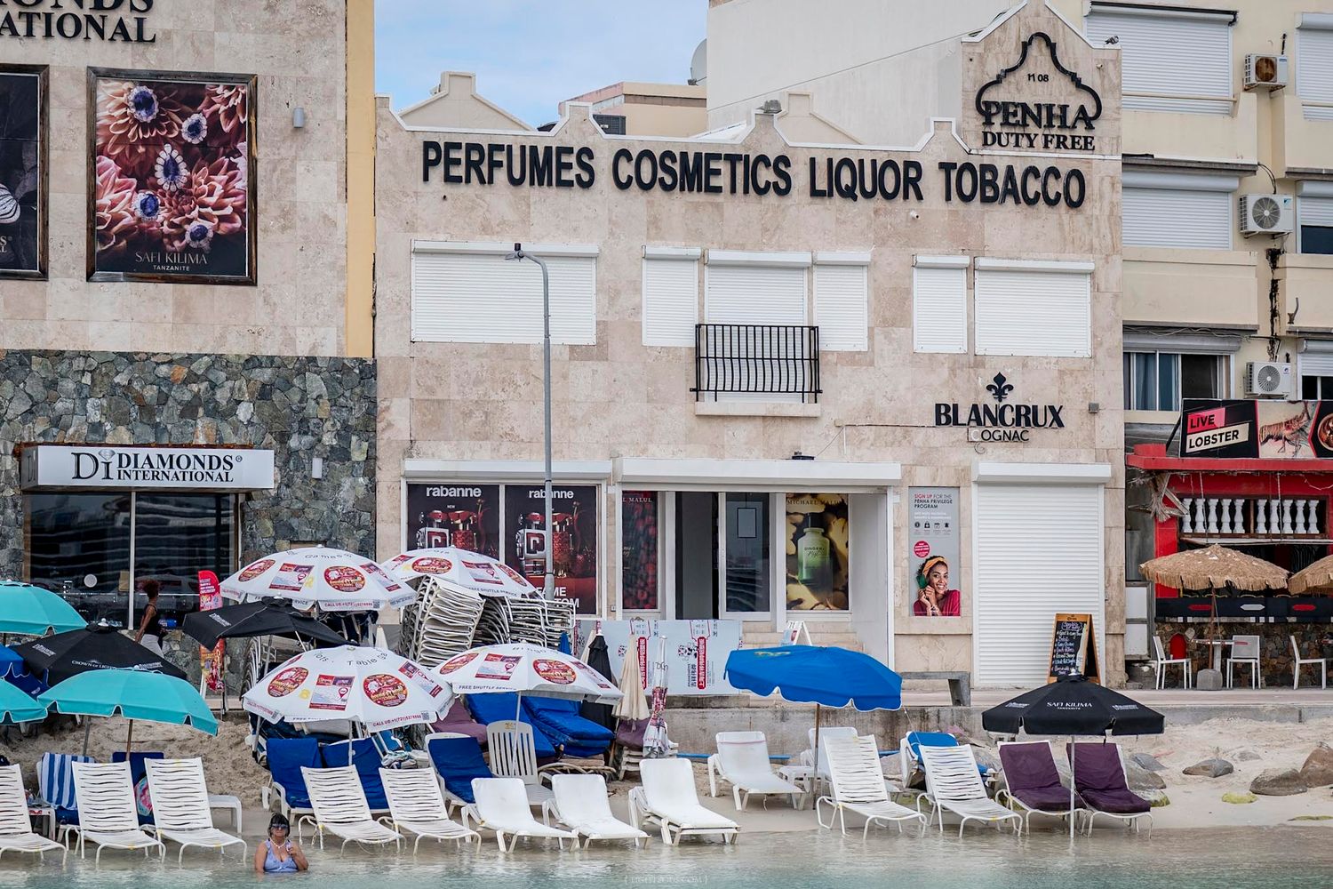 Beachfront duty-free shop selling perfumes, cosmetics, liquor, and tobacco with beach chairs.