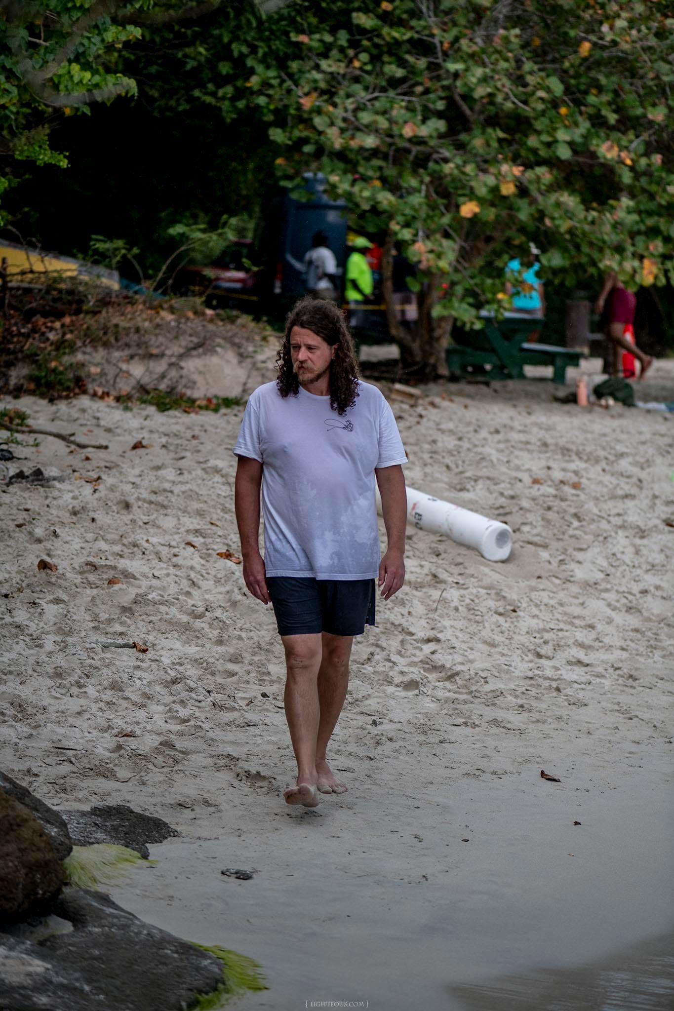 Long-haired person in white t-shirt and dark shorts walking barefoot on a sandy beach near water.