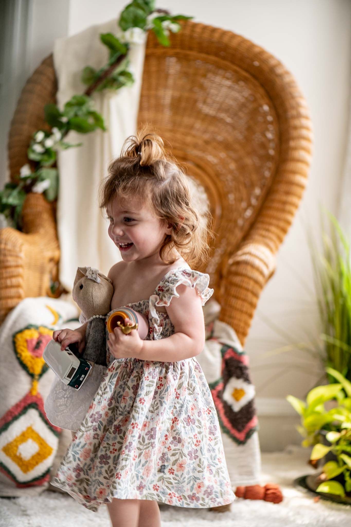 A toddler in a floral dress plays and laughs in front of a wicker peacock chair with colorful decorative elements.