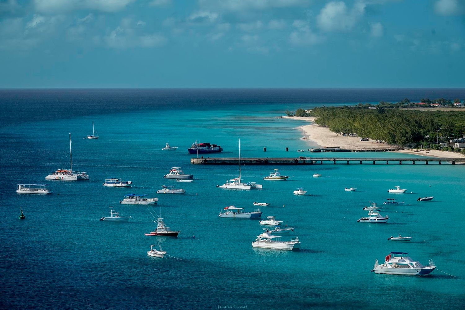 Aerial view of turquoise Caribbean waters with multiple white boats anchored near a tropical island pier and sandy beach.