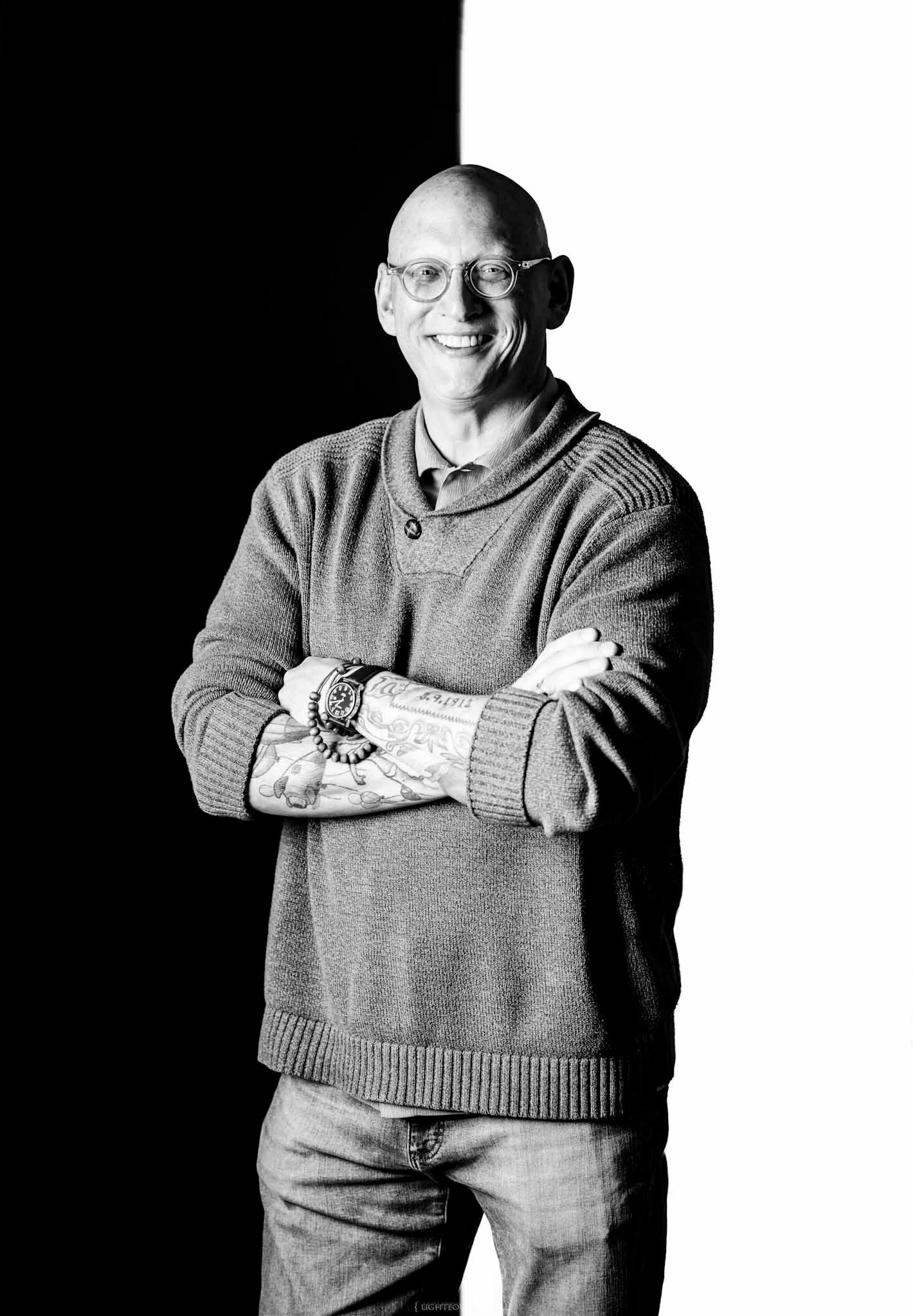 Bald man with glasses and tattoos smiling, arms crossed, wearing a sweater, in a black and white portrait photo.