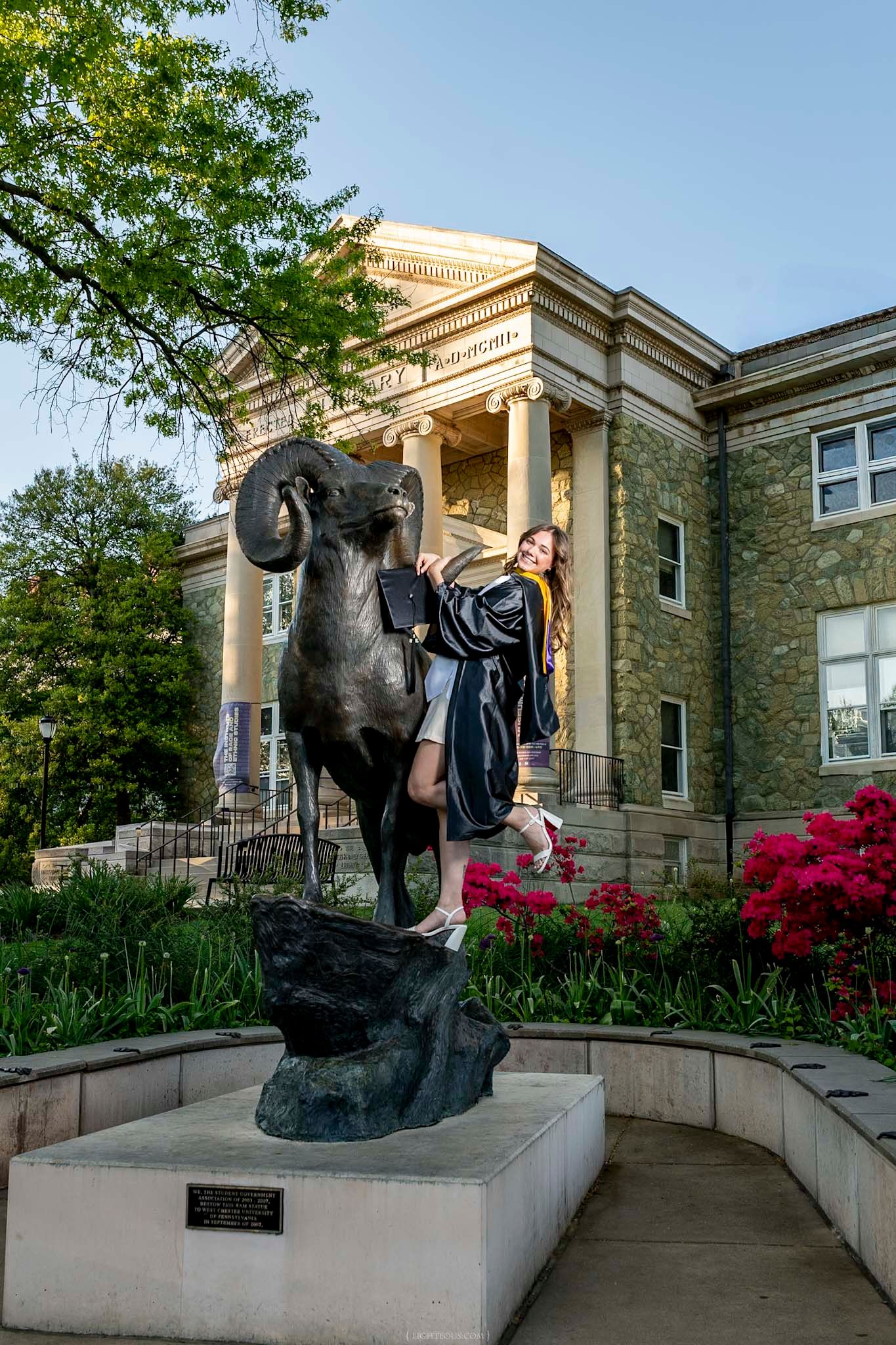 Graduate in cap and gown posing with bronze ram statue outside historic stone university building with red flowers.