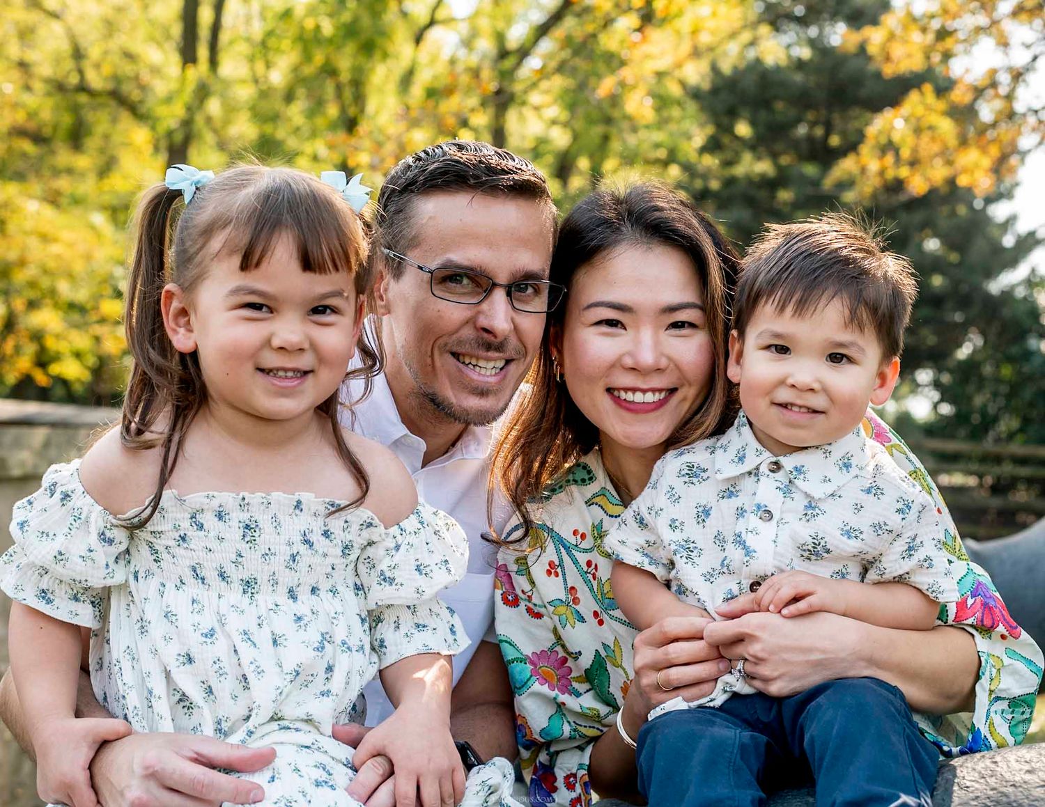 A happy family of four smiling outdoors in autumn, parents holding two young children in matching floral outfits.
