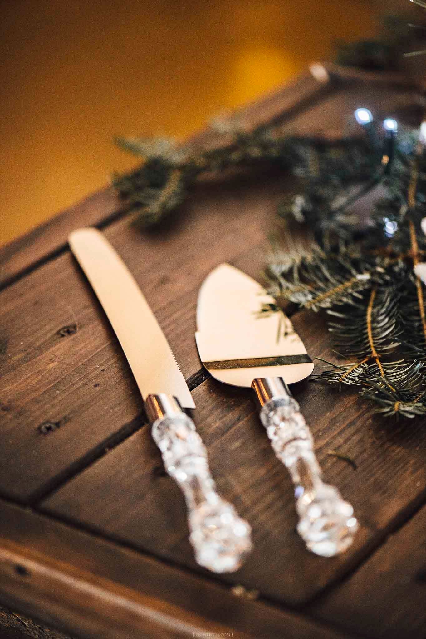 Gold cake knife and server set with crystal handles on rustic wood table with pine branches.