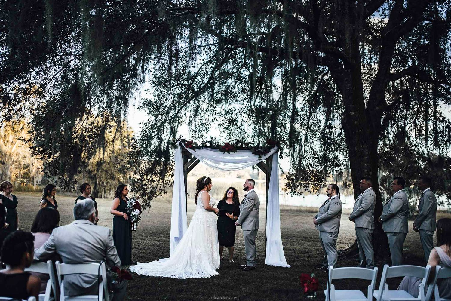 Outdoor wedding ceremony under a large oak tree with bride, groom, and officiant at a rustic arch altar.