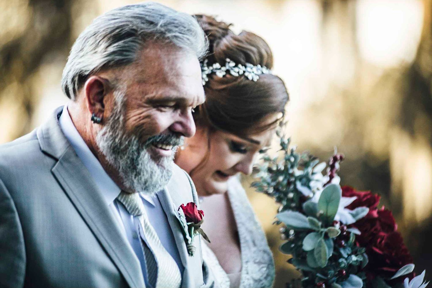 Father and bride share a joyful moment, man in gray suit with red boutonniere, bride holding burgundy bouquet.