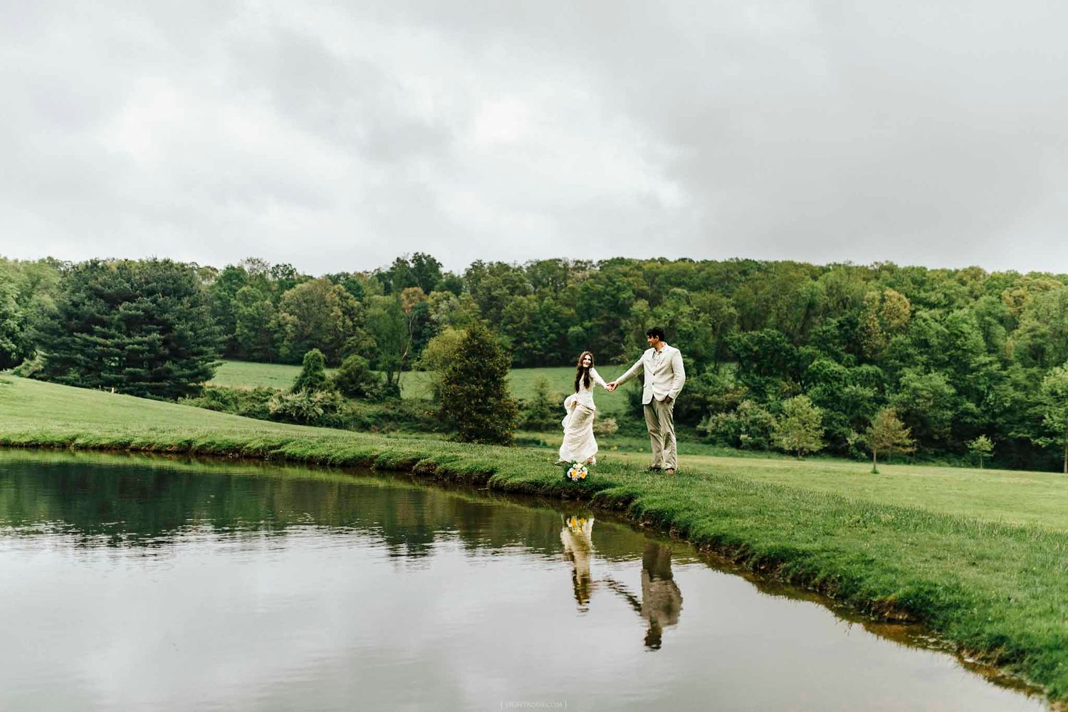Couple holding hands walking along a serene pond with lush green hills and trees reflected in the water.