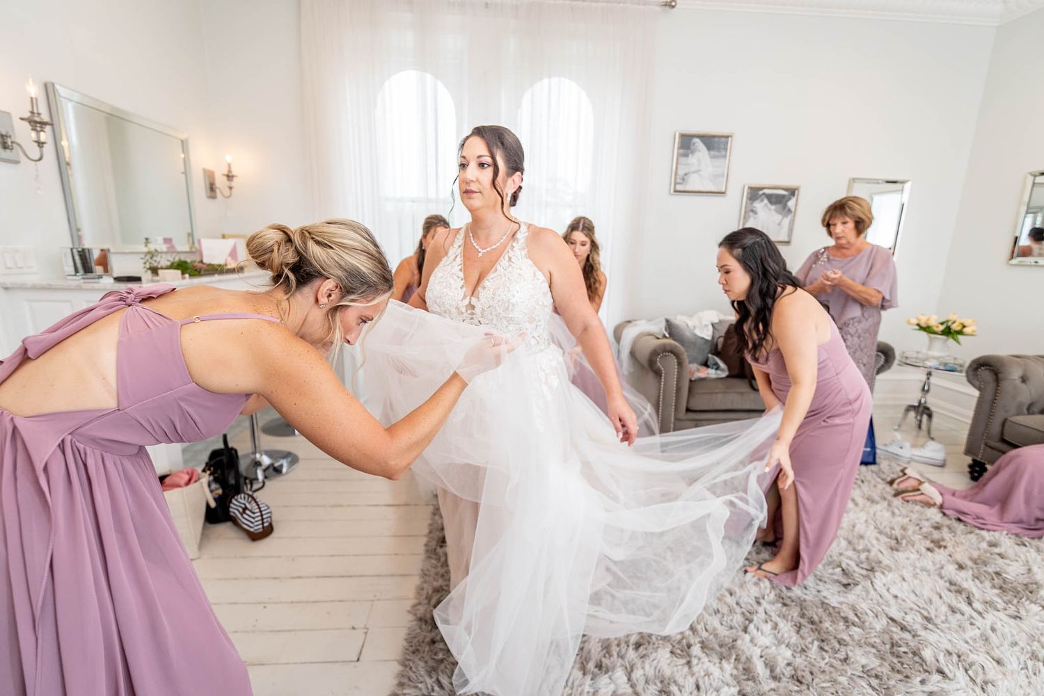 Bridesmaids in lavender gowns help a smiling bride arrange her white wedding dress in an elegant bridal suite.