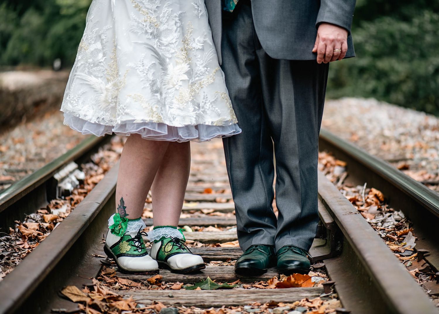 Bride and groom standing on railroad tracks, bride wearing white dress and green sneakers, groom in gray suit.
