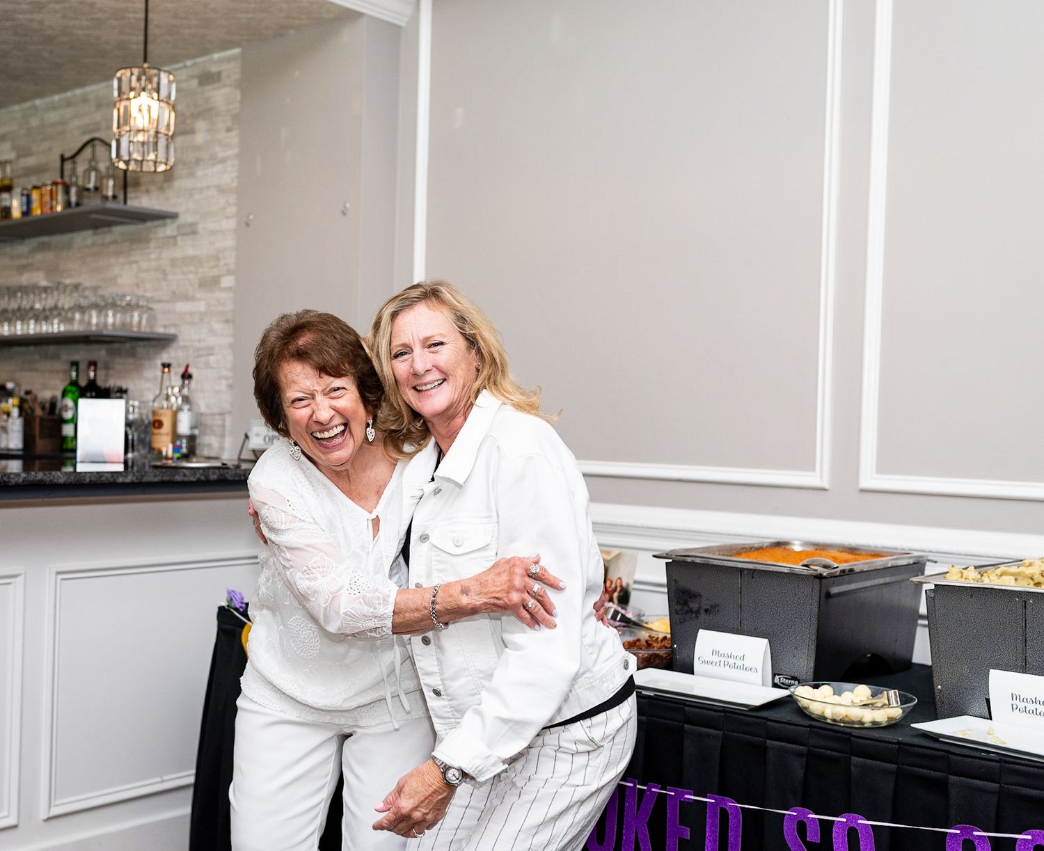 Two smiling women in white outfits embracing at an indoor party near a bar and food table.