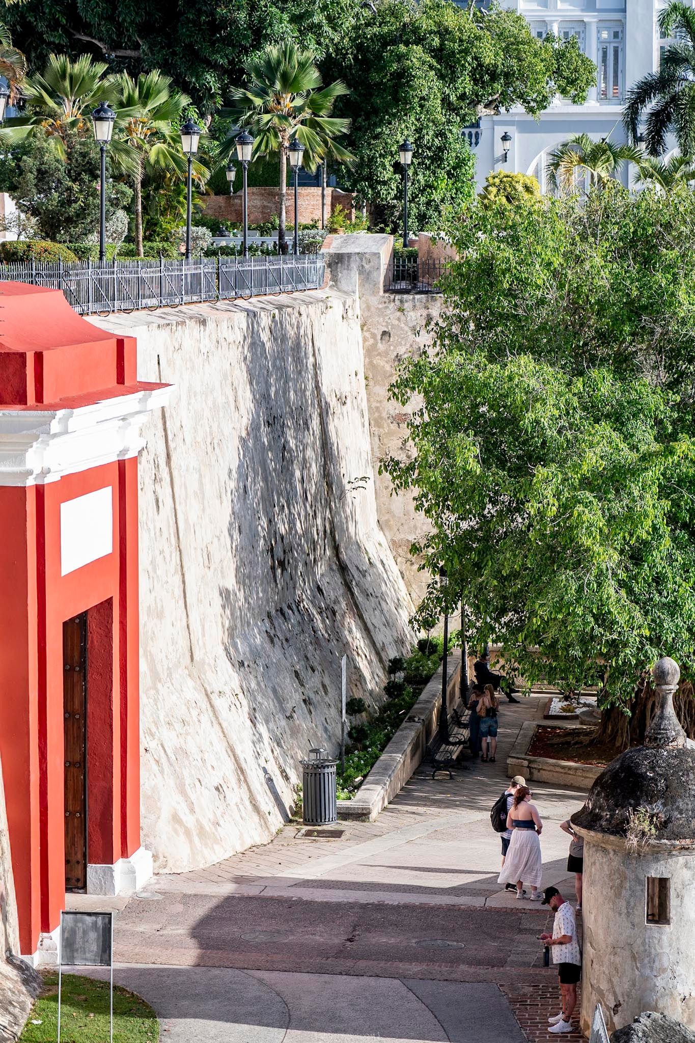 Historic stone fortress wall in Old San Juan, Puerto Rico, with tourists walking along a tree-lined path beside a red building.