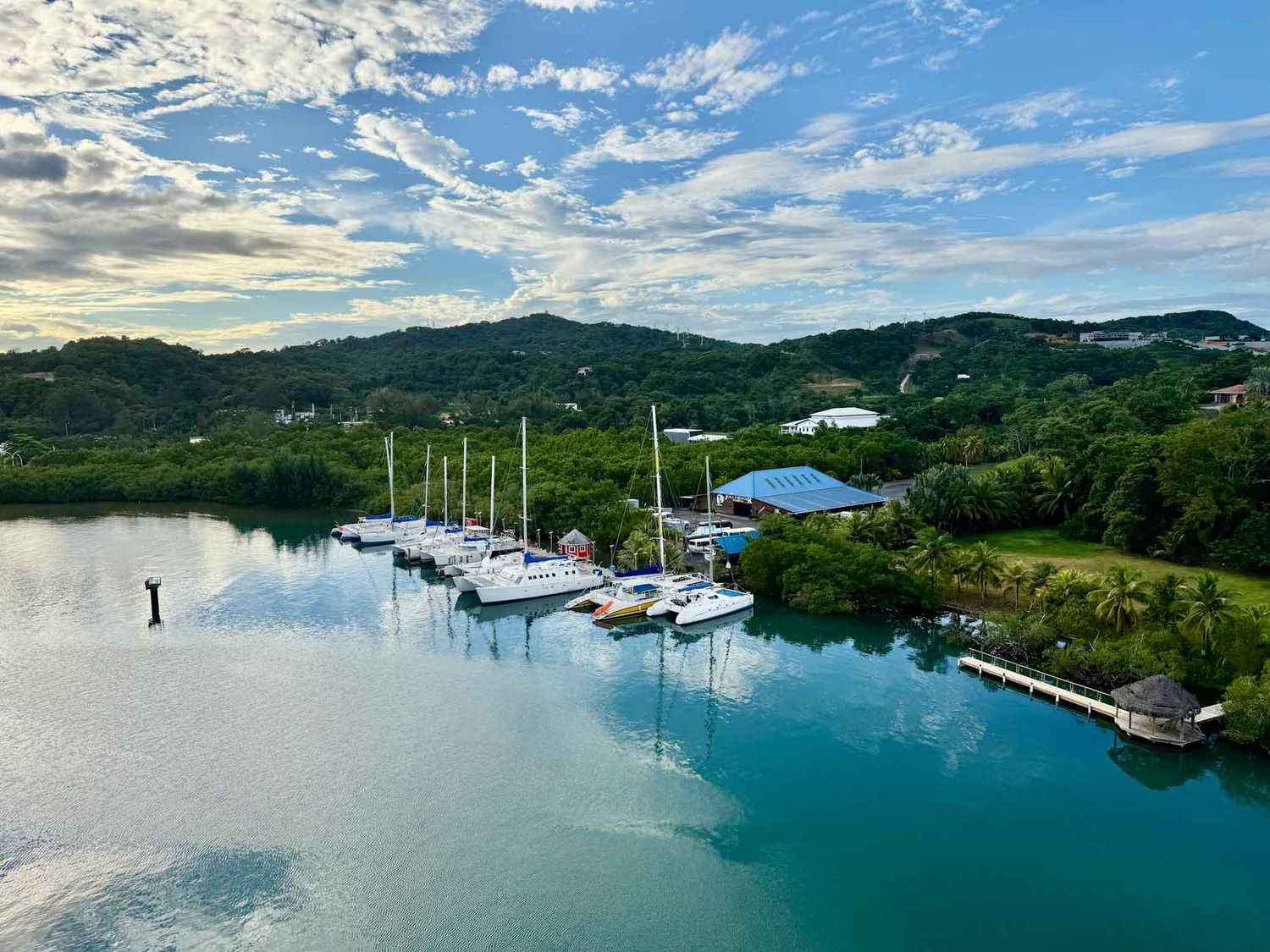 Sailboats docked at a tranquil marina surrounded by lush green hills and calm turquoise waters under wispy clouds.