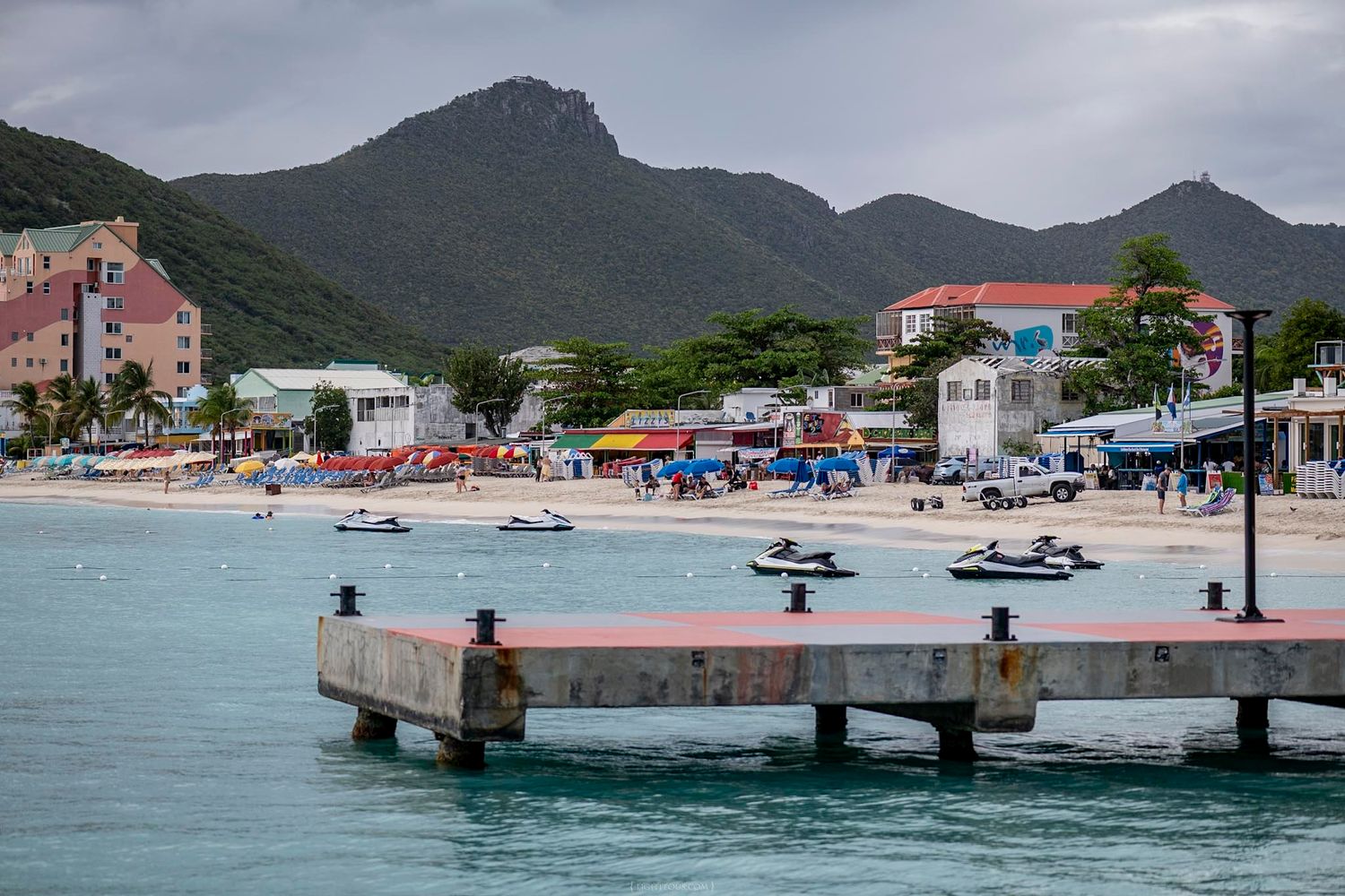 Jet skis on turquoise Caribbean waters near a pier with colorful beachfront buildings and lush green mountains in the background.
