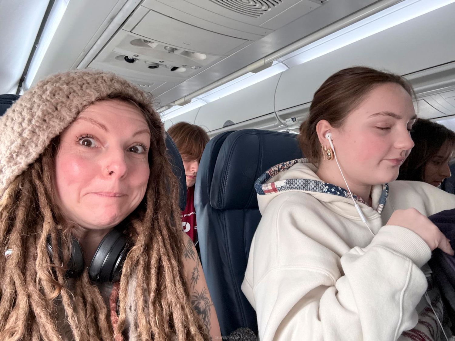 Two women sitting in airplane seats, one with dreadlocks and headphones, another wearing earbuds and a hoodie.