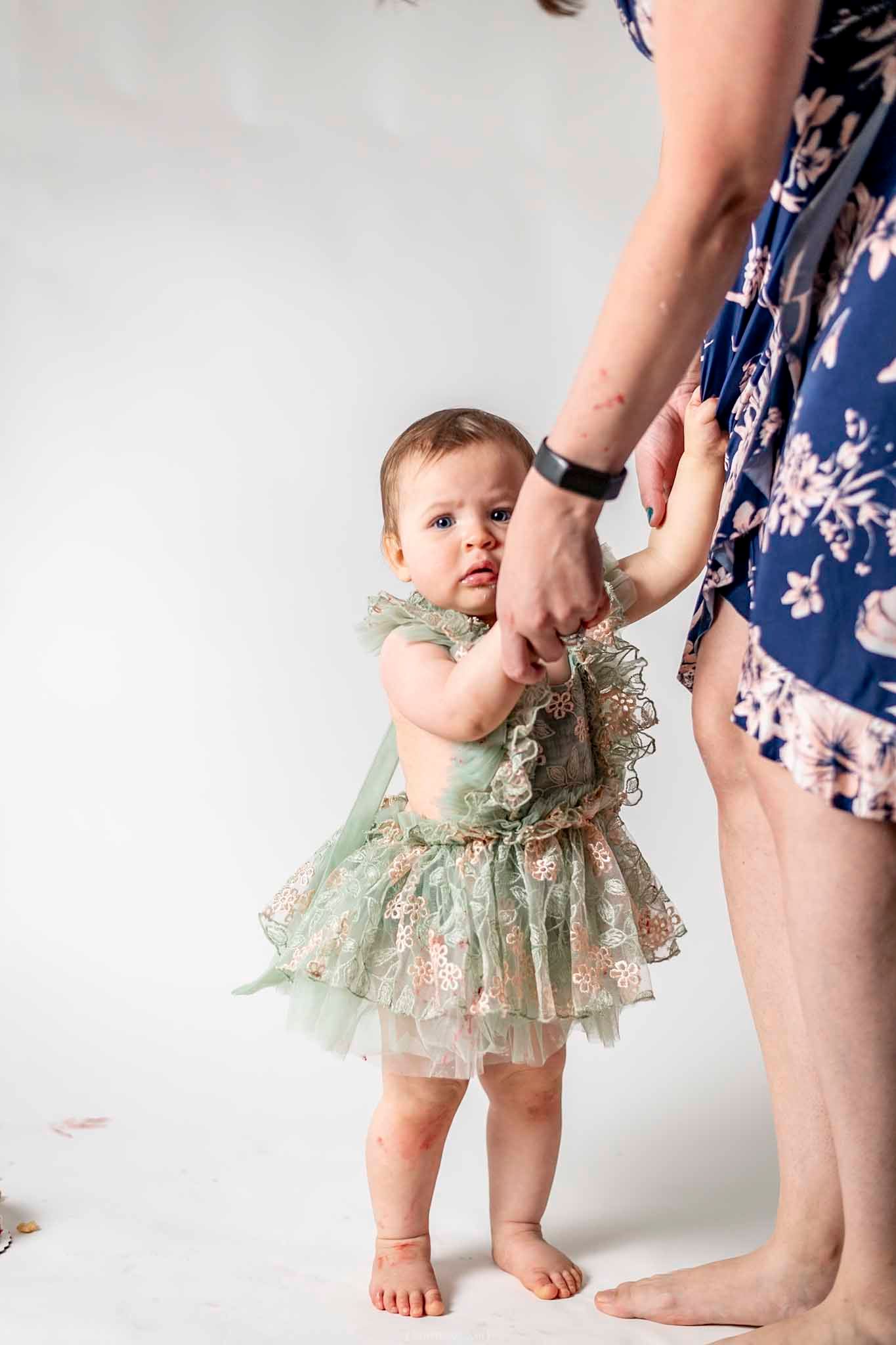 A baby girl in a green floral tutu dress holds an adult's hands while learning to stand on a white studio background.