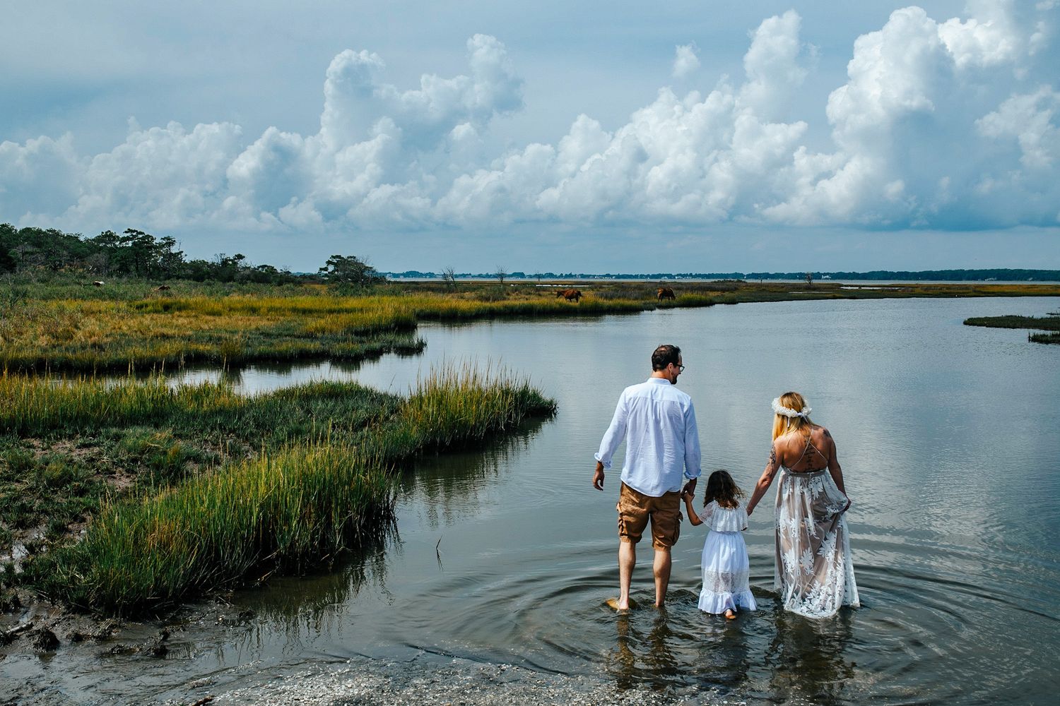 Family of three wading in a coastal marsh, holding hands, with lush wetlands and dramatic clouds in the background.