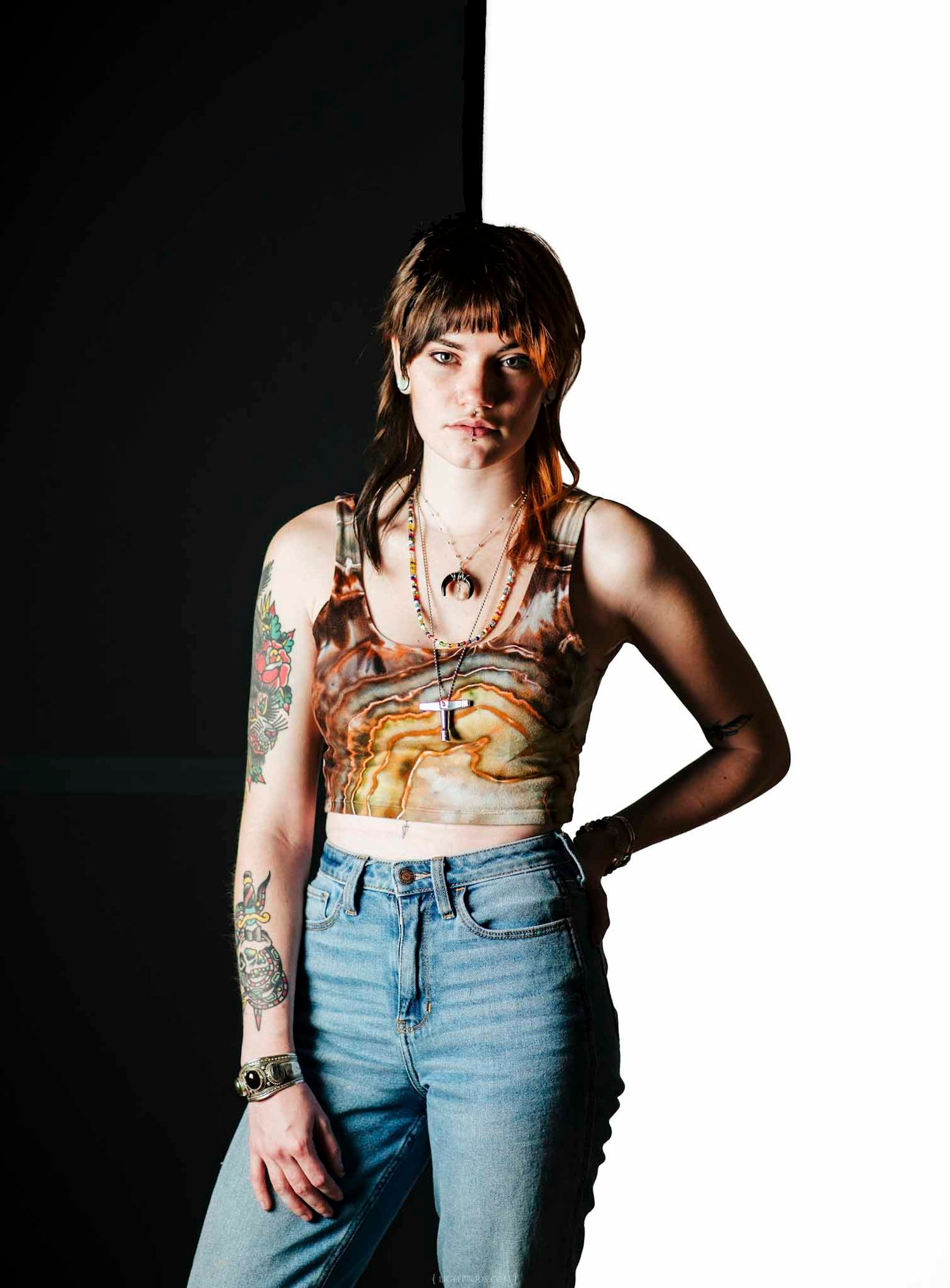 Young woman with tattoos wearing a crop top and jeans poses confidently against a black and white background.
