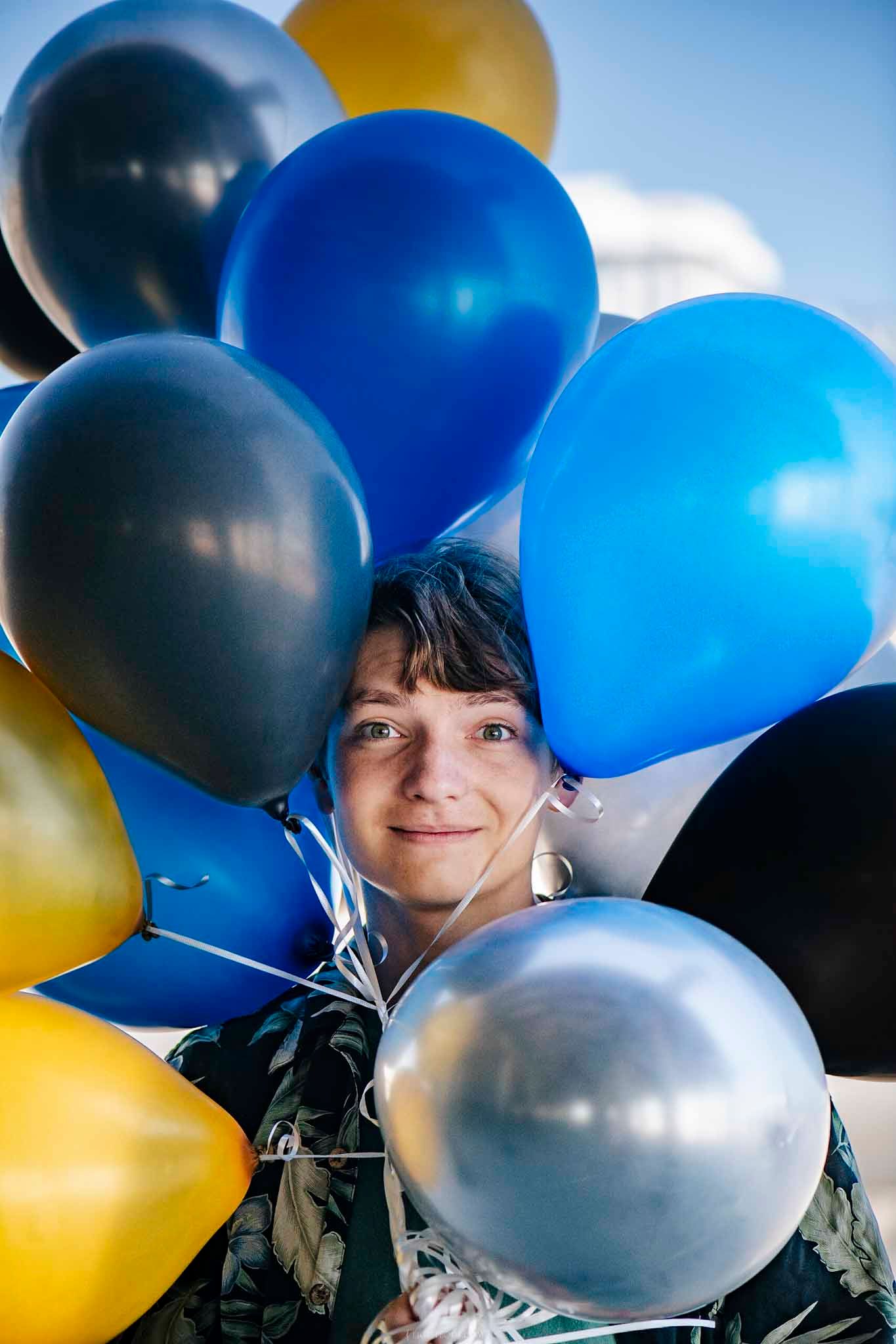 Smiling woman surrounded by colorful blue, black, silver, and yellow balloons outdoors.