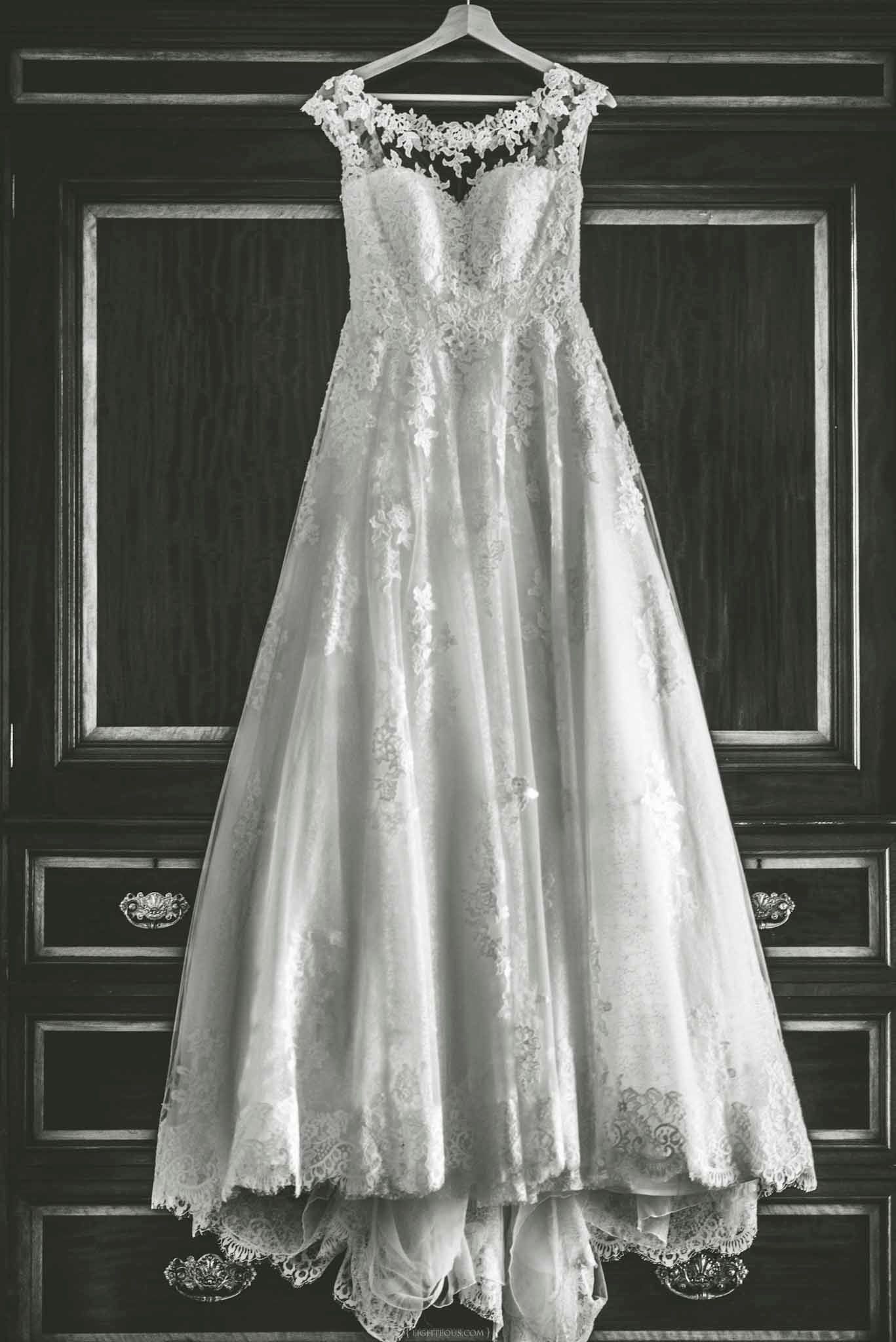 Elegant lace wedding dress with cap sleeves and full skirt hanging on wooden armoire in black and white photo.