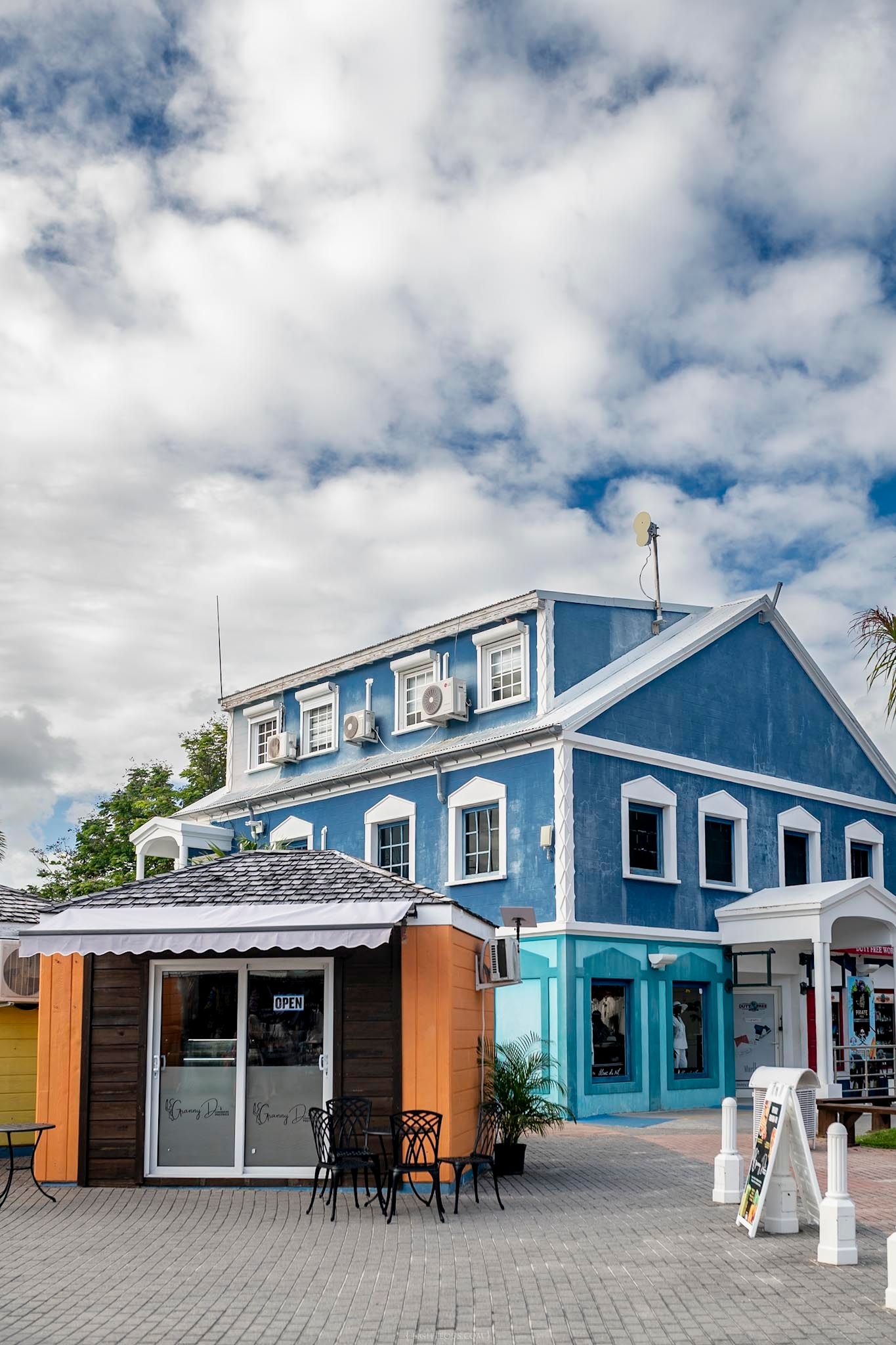 Colorful blue Caribbean building with white trim and outdoor seating on a brick plaza under a cloudy sky.