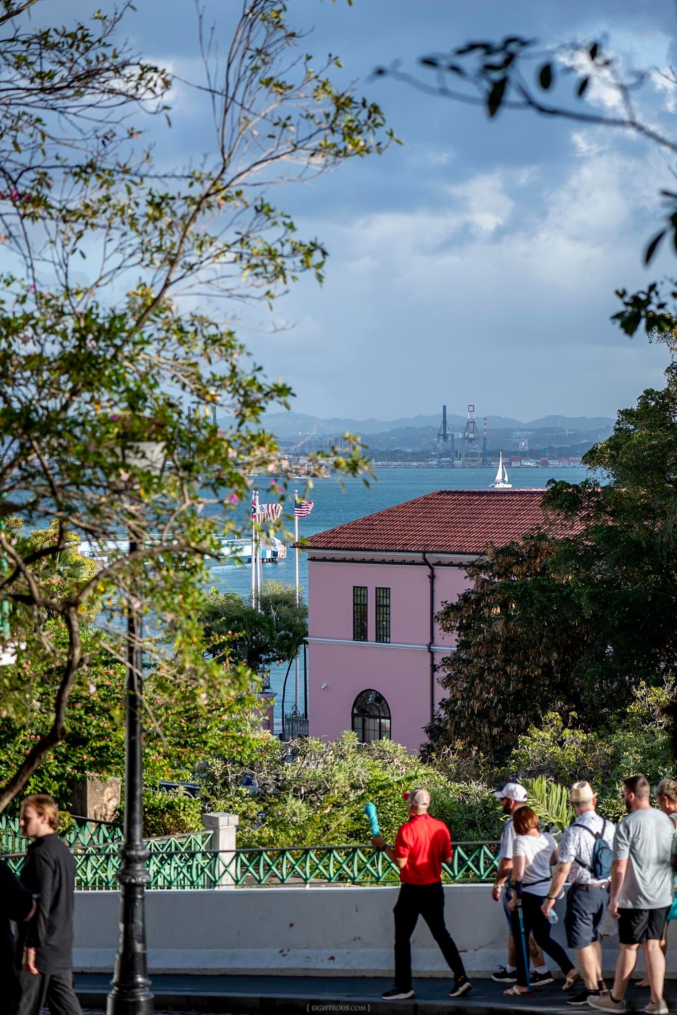 Pedestrians walk along a scenic overlook path with palm trees, a pink building, and a distant bridge over blue water.