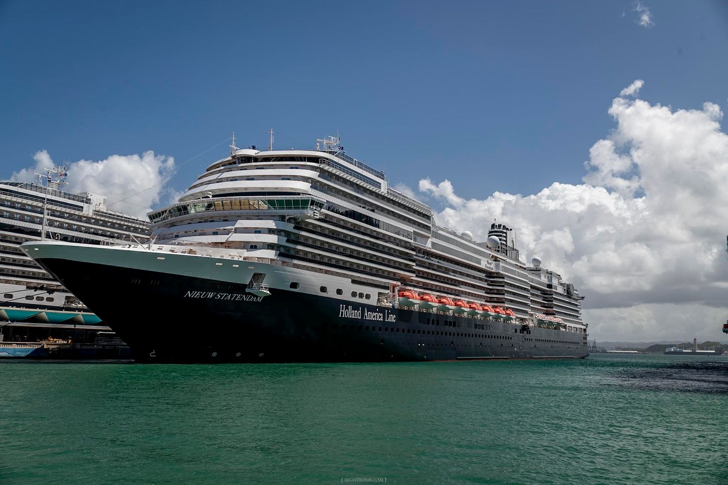Two large Holland America Line cruise ships docked at a tropical port under partly cloudy skies.