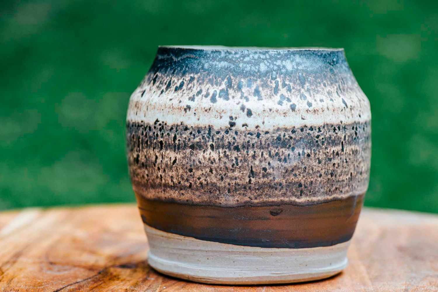 Handcrafted ceramic vase with layered brown, white, and blue glaze sitting on a wooden surface with green background.